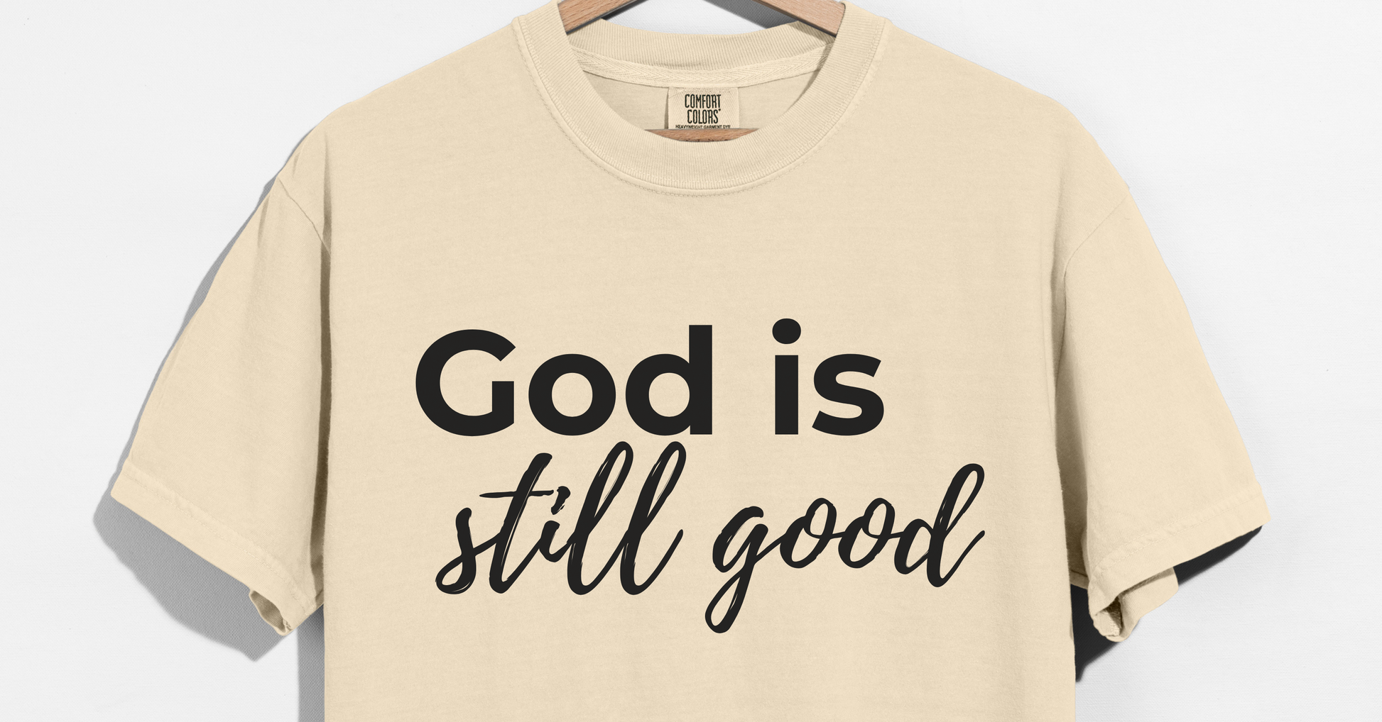 Beige t-shirt with 'God is still good' text on a hanger against a white background